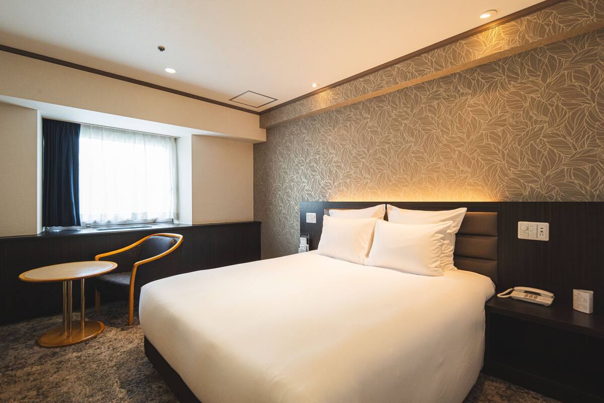 Habitación de hotel con una cama grande, una mesita redonda pequeña, una silla, una mesita de noche con un teléfono y una ventana con cortinas.