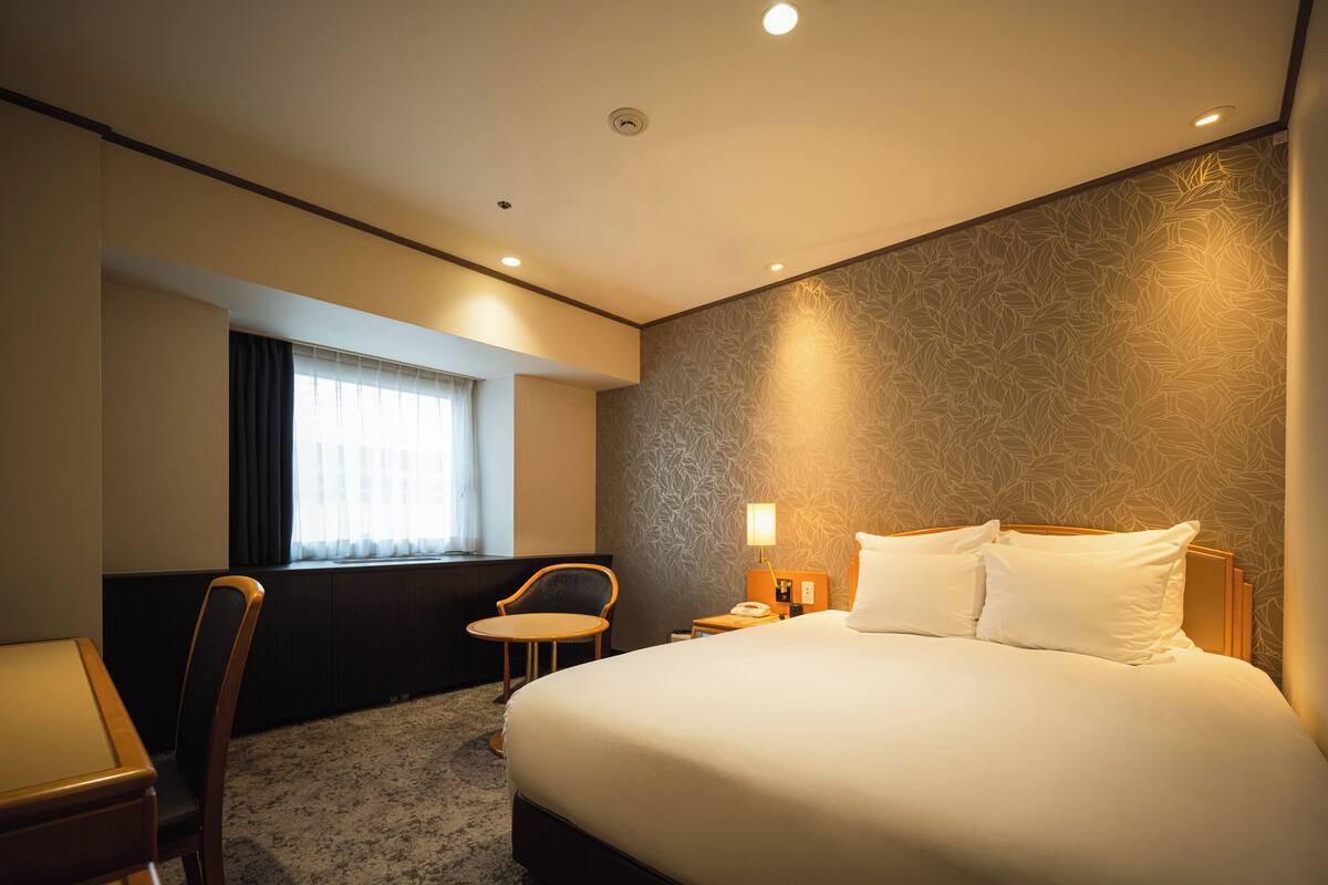 Habitación de hotel con una cama grande, una mesita pequeña, una silla, un ventanal con cortinas, una pared con diseño de hojas y una mesita de noche con lámpara.