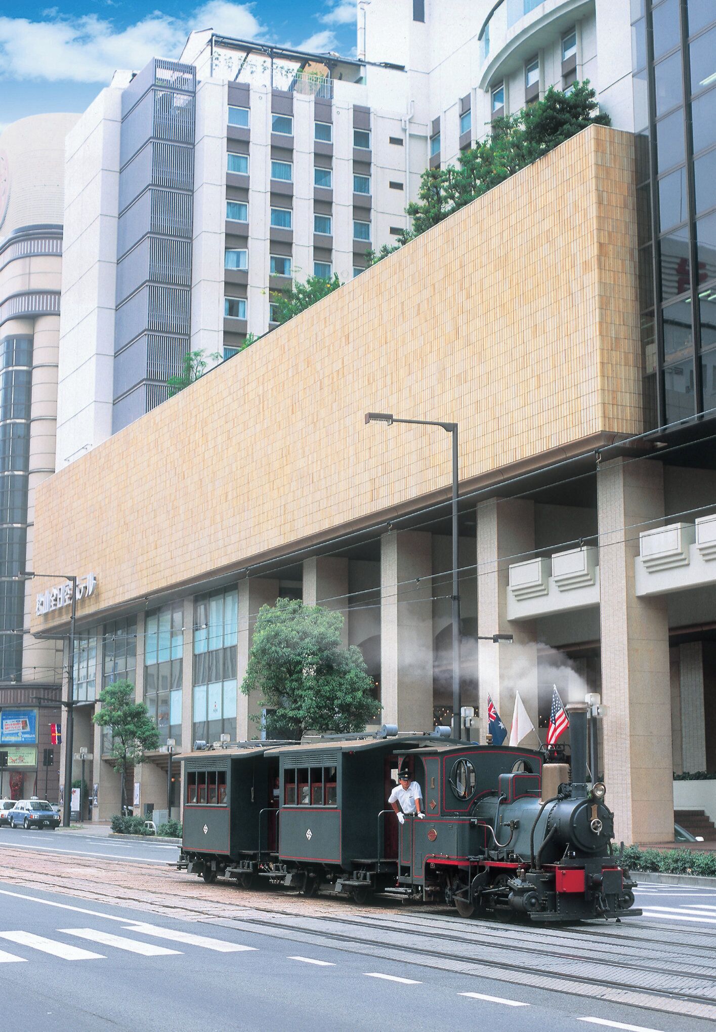 A steam train with a conductor, passing by a modern building with large windows and a flat roof.