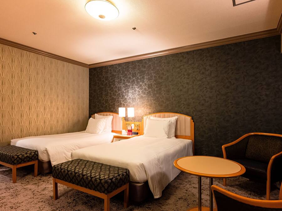 A hotel room with two beds, a small table, a chair, and a bench.