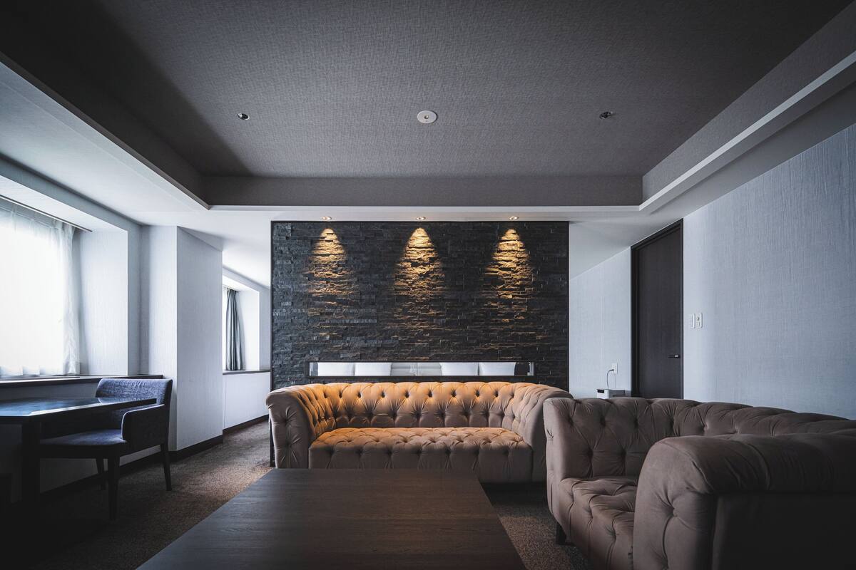 Una sala de estar moderna con un sofá capitonado, una mesa de comedor y sillas. Hay un ventanal grande a la izquierda y una pared de piedra con iluminación empotrada.
