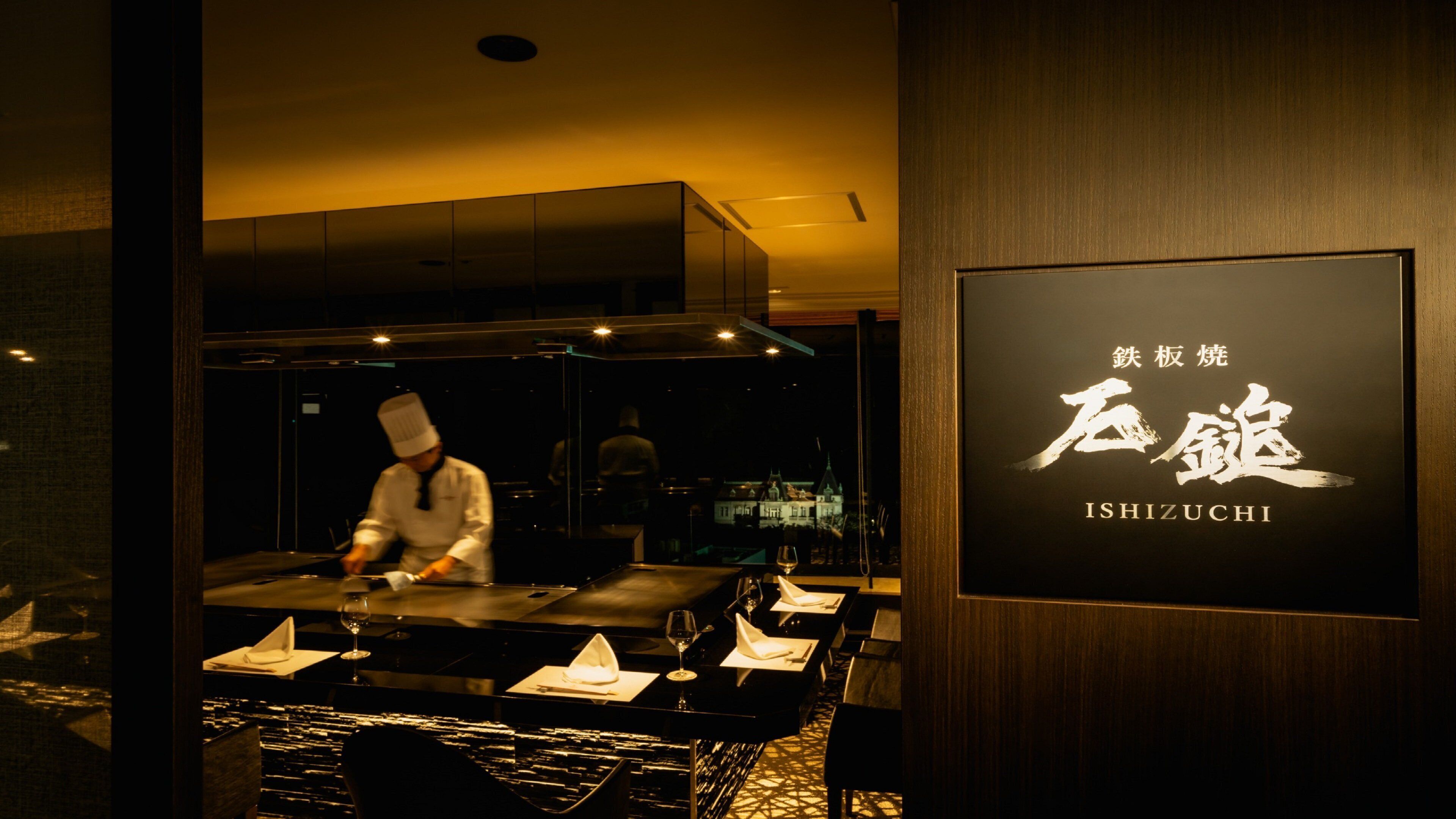 L’intérieur d’un restaurant où l’on voit un chef préparer des plats, une table dressée avec des verres et des serviettes, et une enseigne avec des caractères japonais.