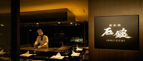 L’intérieur d’un restaurant où l’on voit un chef préparer des plats, une table dressée avec des verres et des serviettes, et une enseigne avec des caractères japonais.