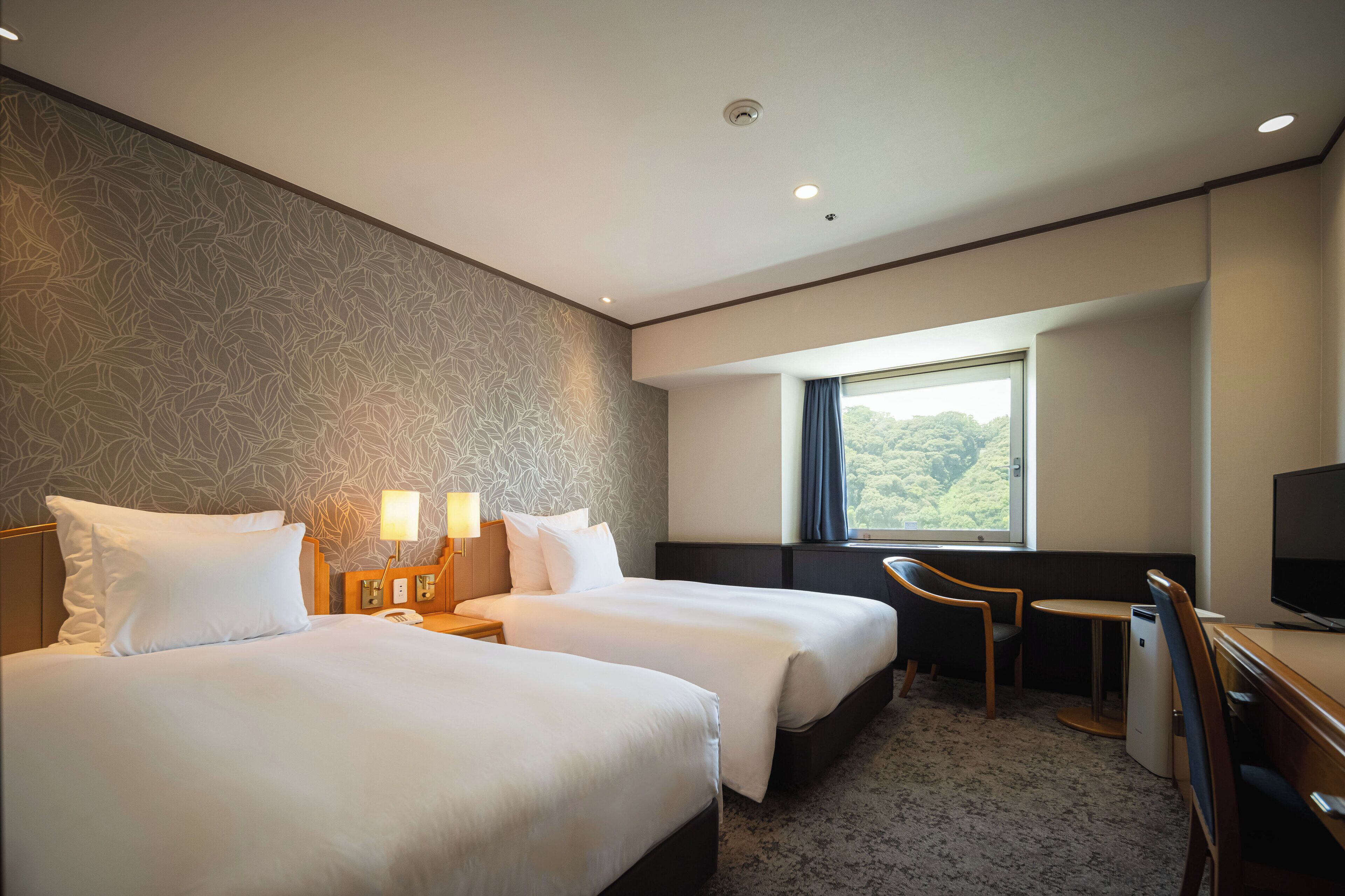 Quarto de hotel com duas camas, uma escrivaninha, uma cadeira, uma televisão e janela com vista para o verde.