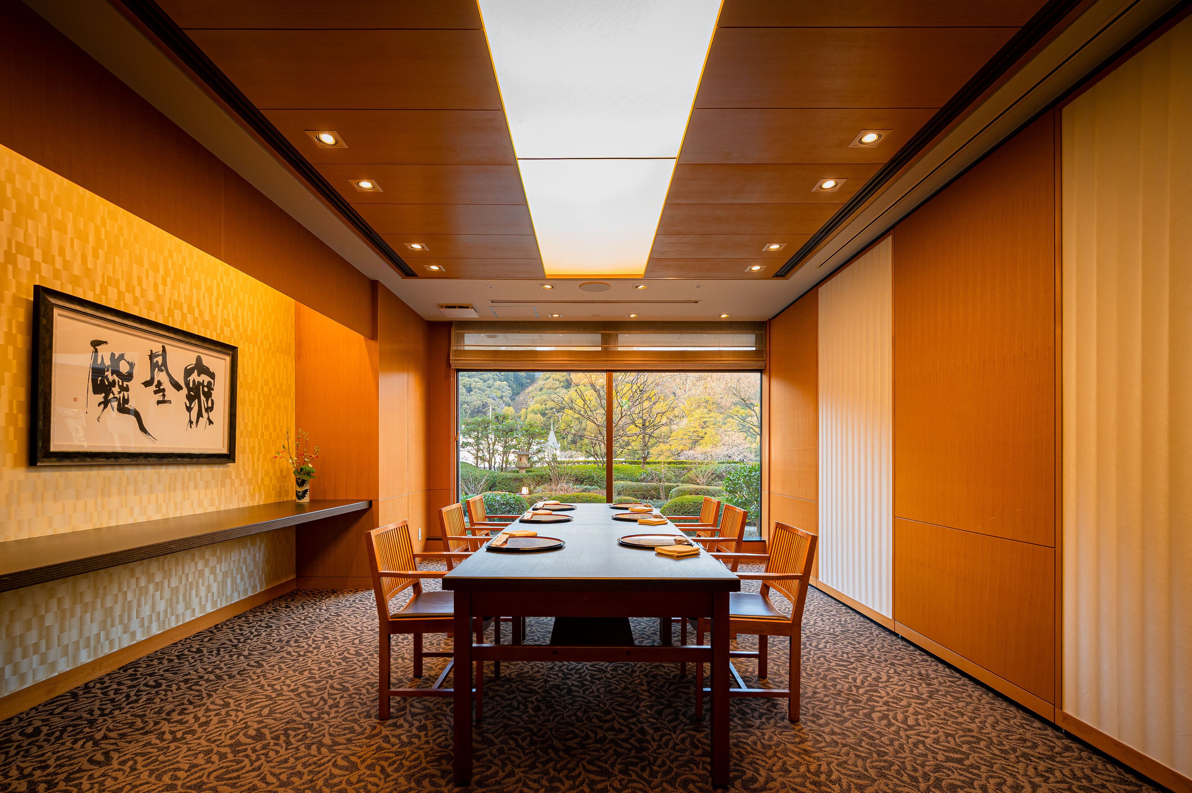 A conference room with a large table, chairs, and a view of trees through large windows.