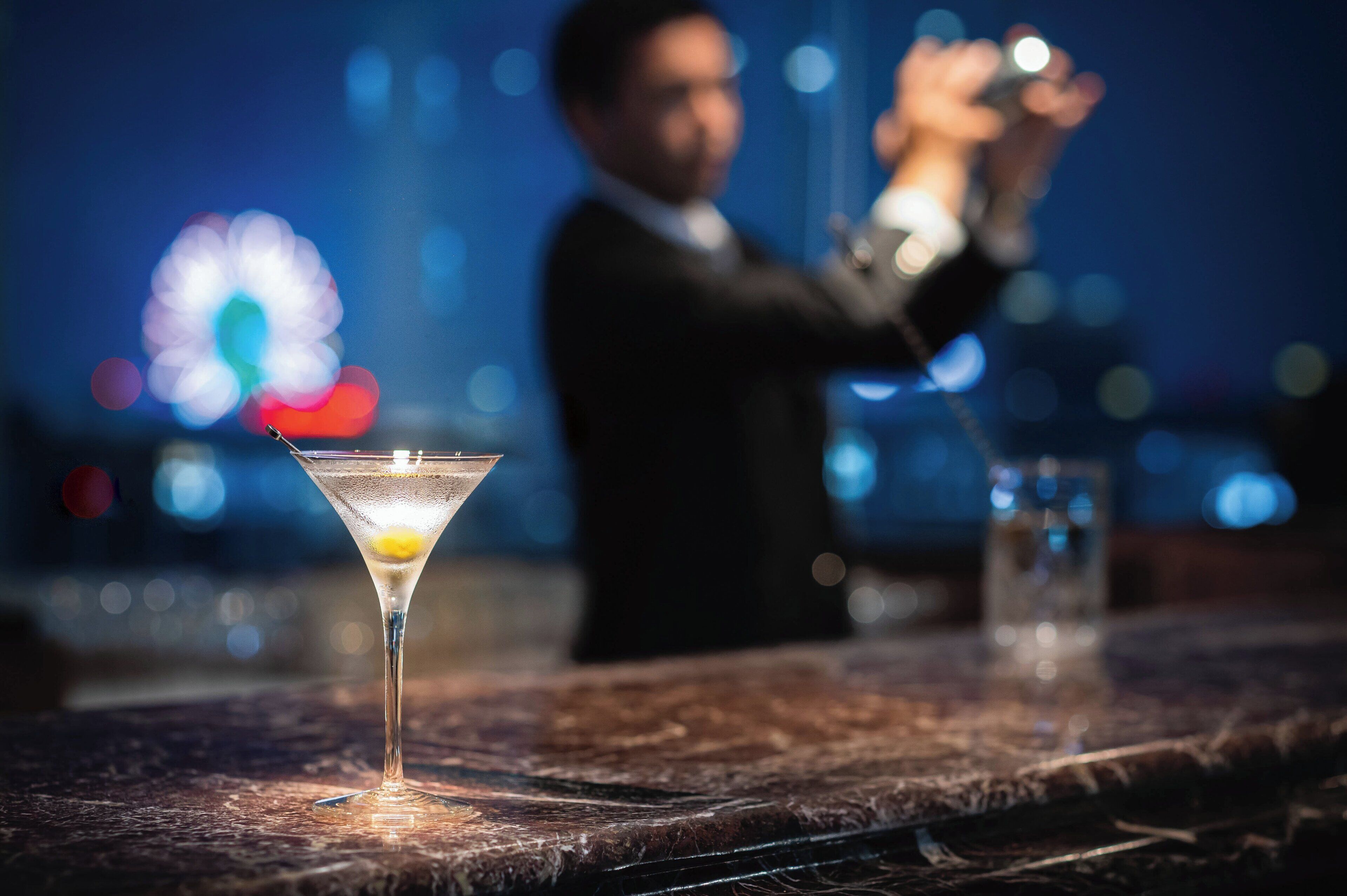Una copa de martini sobre una barra con un fondo borroso de una persona y luces de la ciudad.