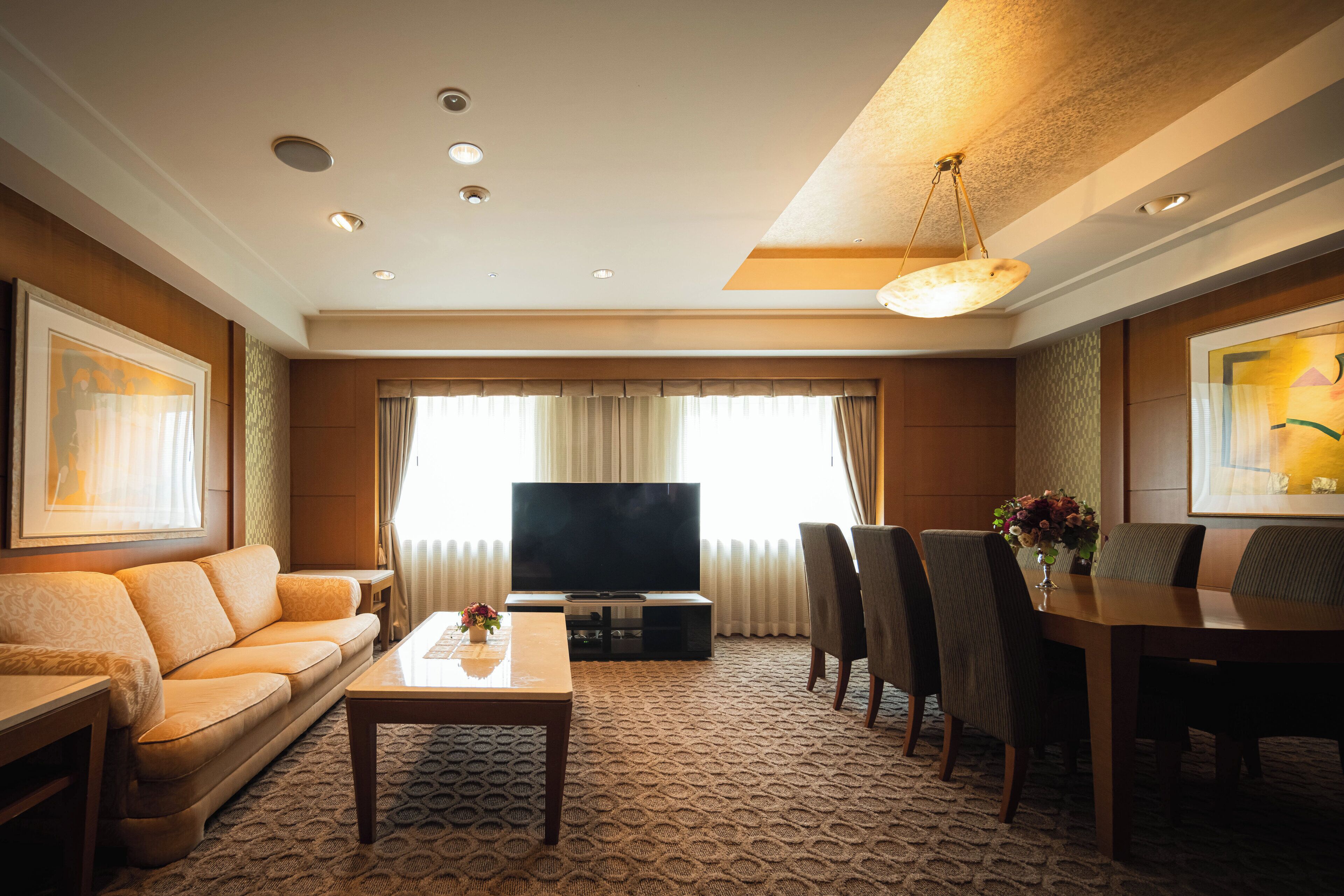 A hotel room with a sofa, TV, dining table, chairs, and a carpeted floor.