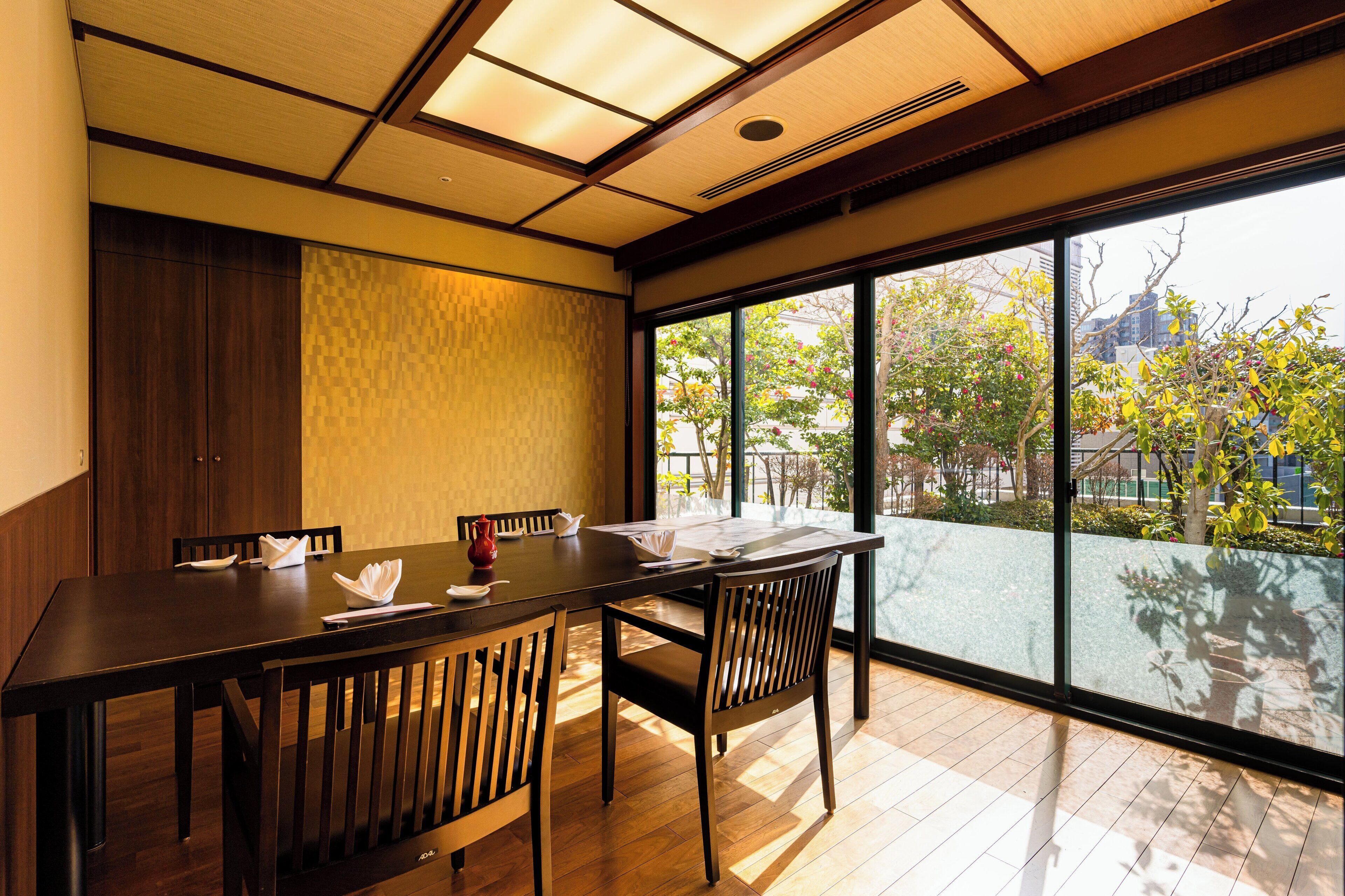 A dining area with a wooden table and chairs, large windows with a view of trees and buildings, and a traditional ceiling design.