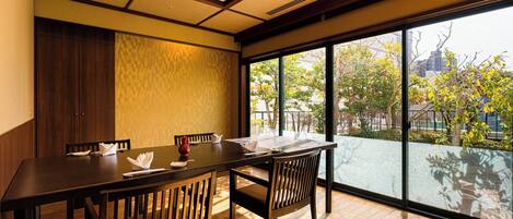 A dining area with a wooden table and chairs, large windows with a view of trees and buildings, and a traditional ceiling design.