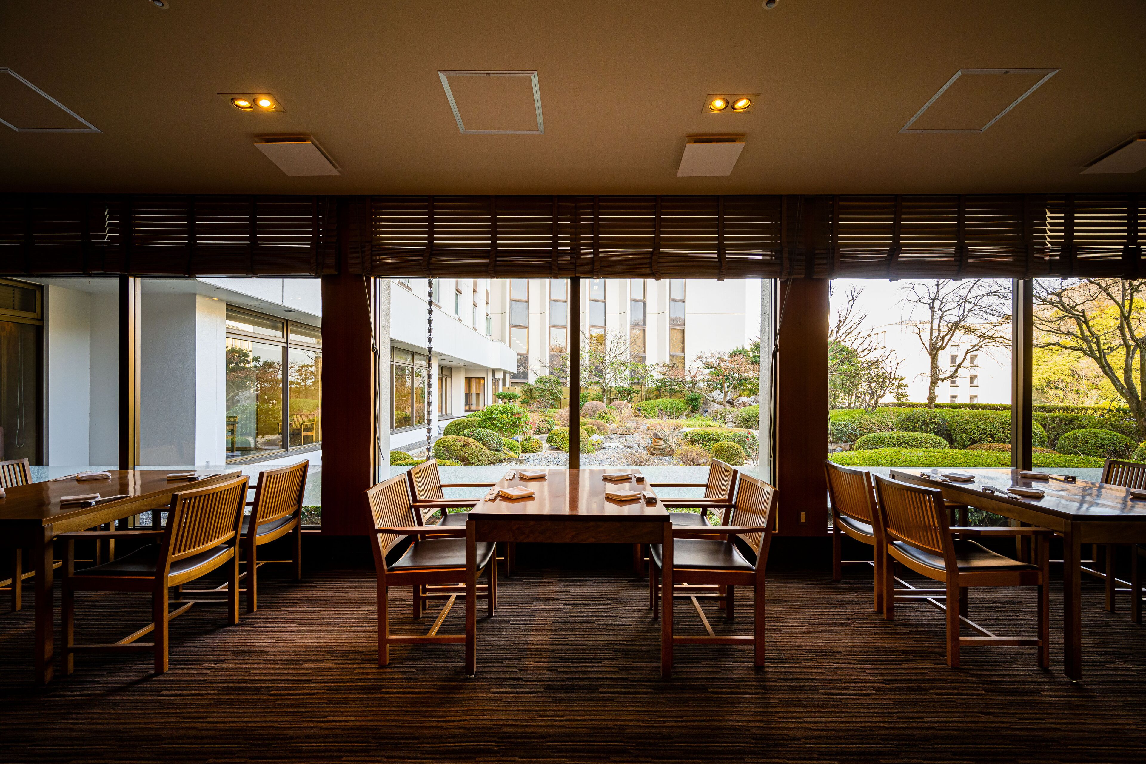 A dining area with wooden tables and chairs, large windows with a view of a garden and buildings.