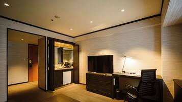 A modern hotel room with a desk, chair, TV, and a small kitchenette.