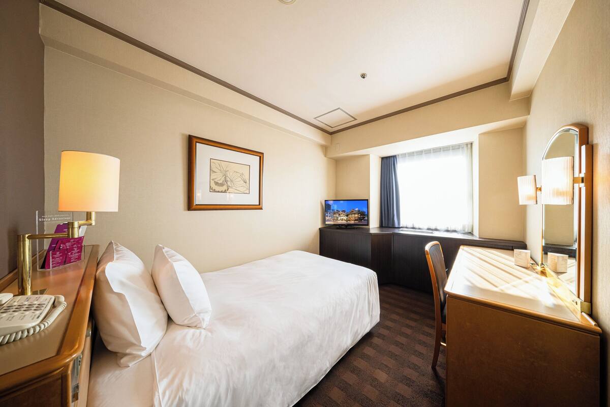 Una habitación de hotel con cama, escritorio, silla, lámpara de escritorio y televisión.