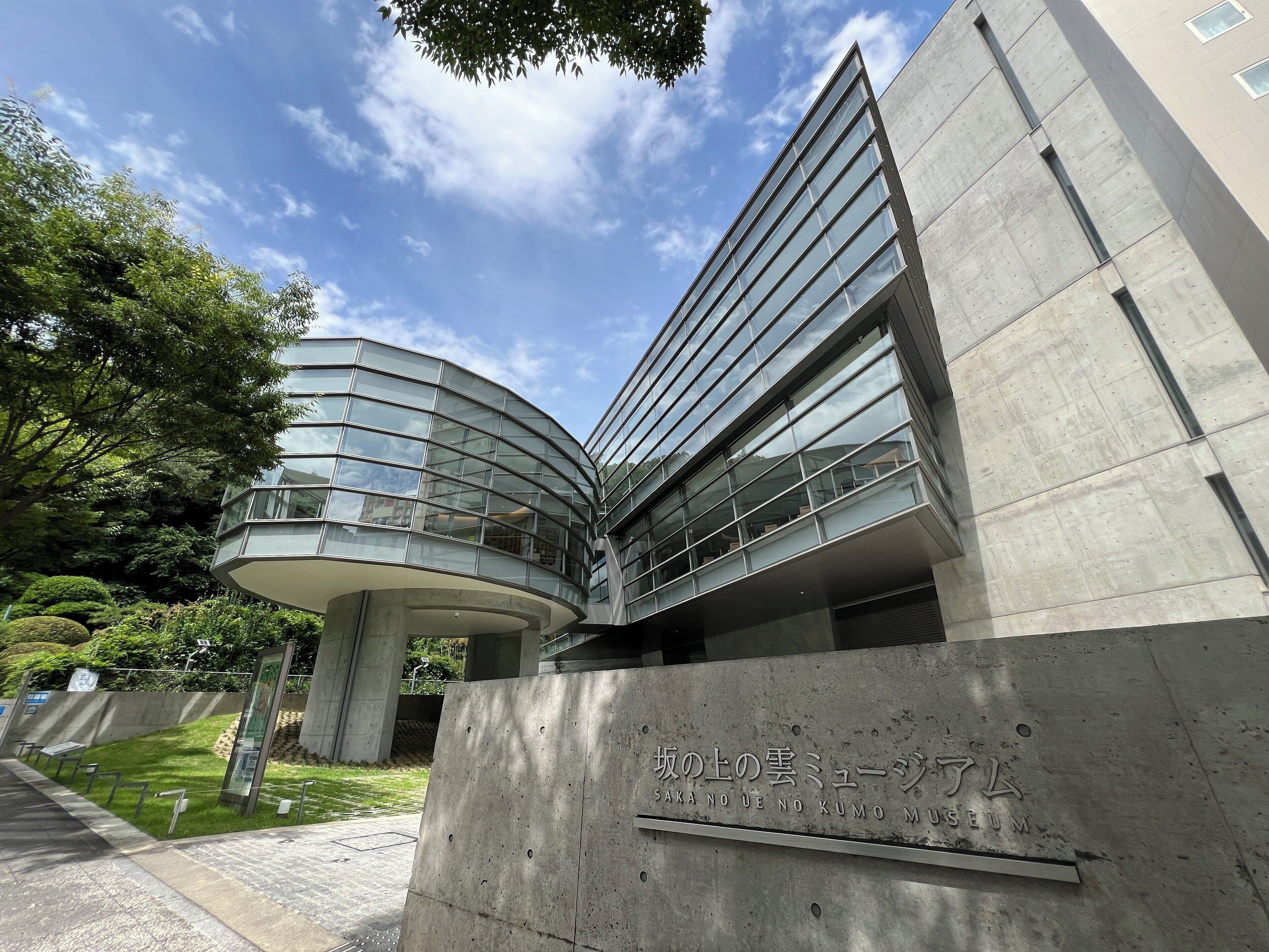 A modern building with a curved glass facade and a sign reading "Saka no ue no Kumo Museum".