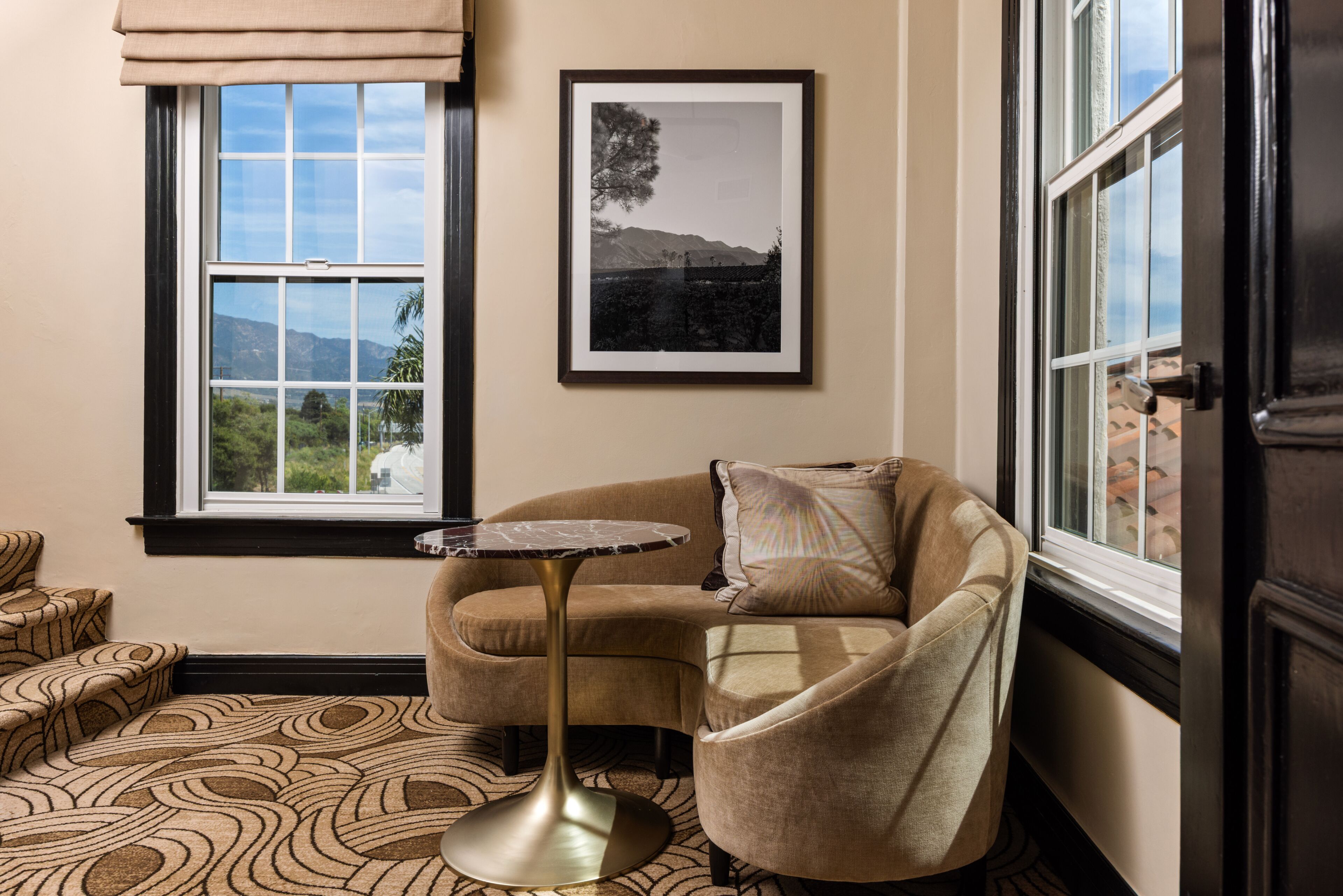 A room with a beige sofa, a small round table, a framed picture on the wall, and a window with a view of greenery and mountains.