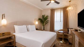 Une chambre d’hôtel comprenant un lit, des tables de chevet, un bureau, une chaise, un ventilateur de plafond, une télévision et une fenêtre avec des rideaux.