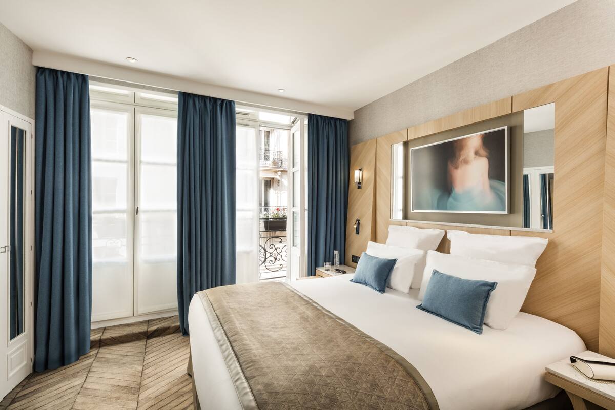 A hotel room with a large bed, a TV mounted on the wall, and a balcony with blue curtains.