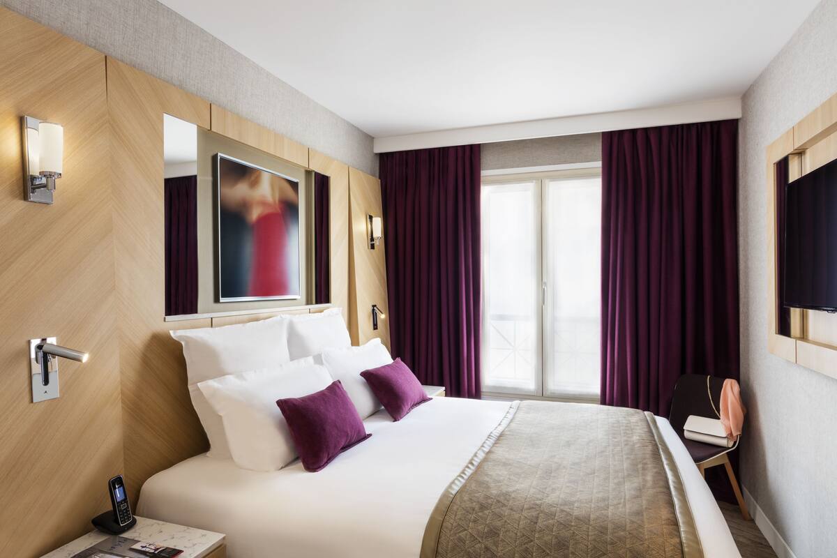 A modern hotel room with a large bed, a bedside table with a phone, a chair, and a TV mounted on the wall.