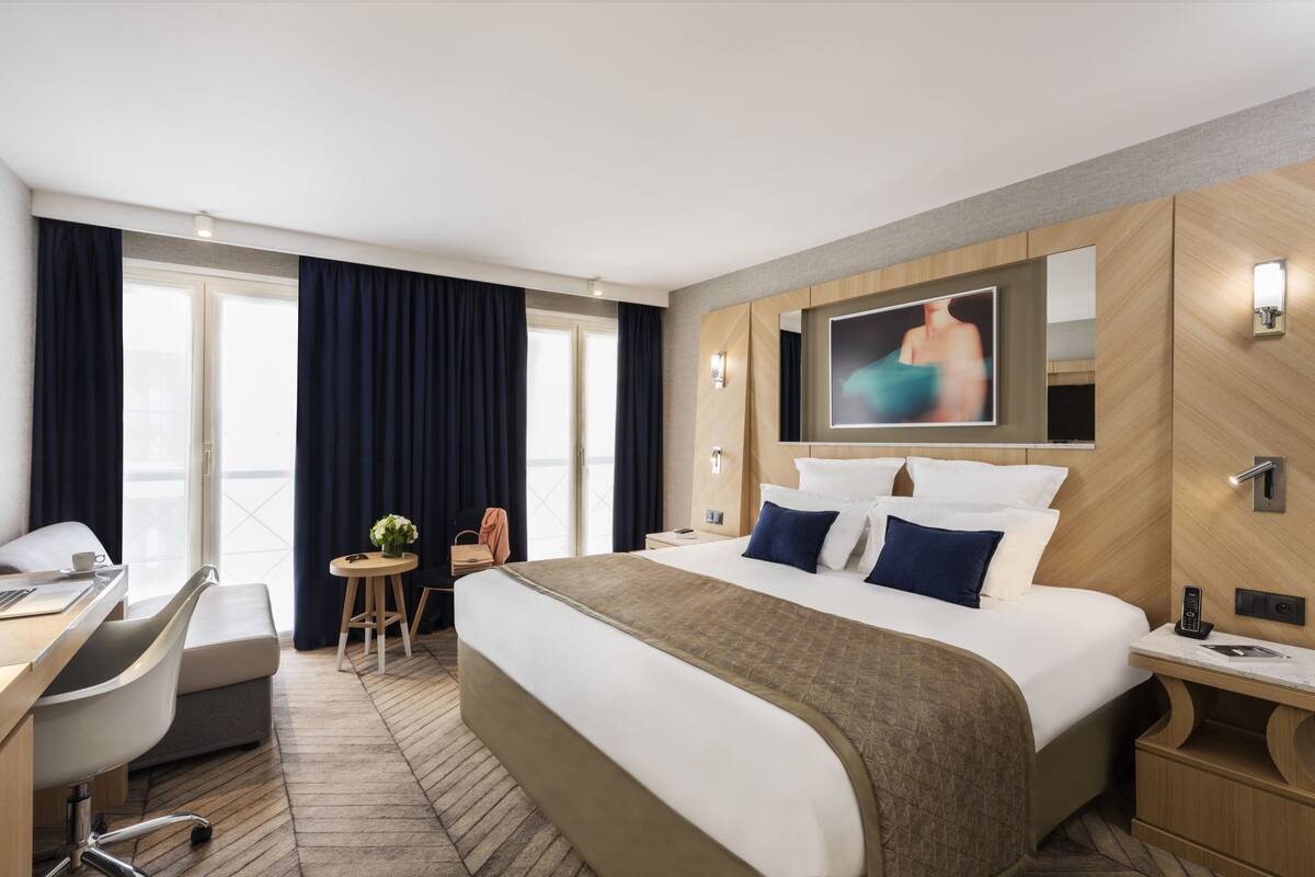 A hotel room with a large bed, a desk, a chair, a small table with a plant, and a TV mounted on the wall.