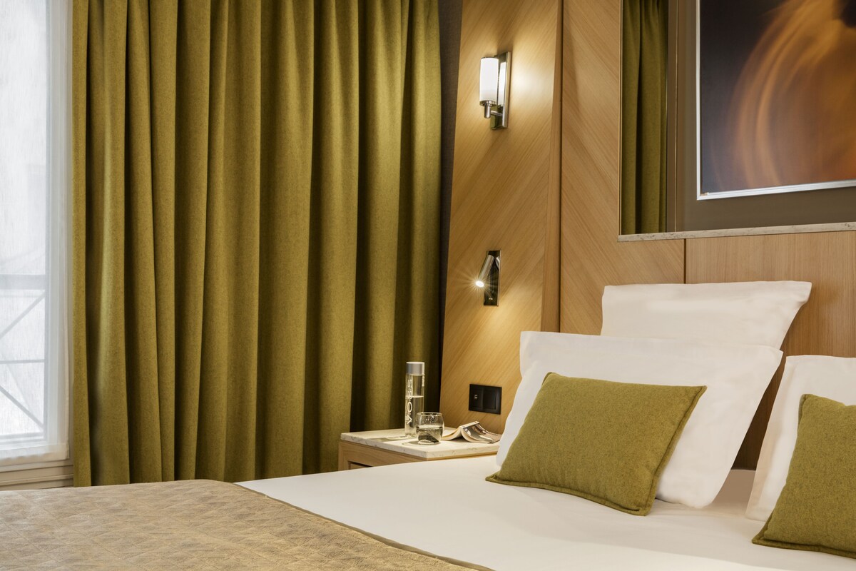 A hotel room with a bed, green curtains, a nightstand with a glass of water, a lamp, and a painting on the wall.