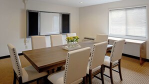 Ein Konferenzraum mit einem Holztisch, sechs weißen Stühlen, einer Whiteboard-Tafel und einem Fenster mit Jalousien.