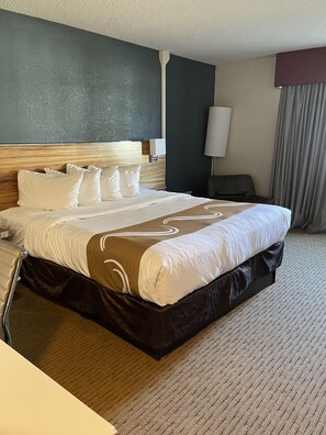 A hotel room with a large bed, a wooden headboard, a bedside table, a lamp, and a view of the outside through a window with curtains.