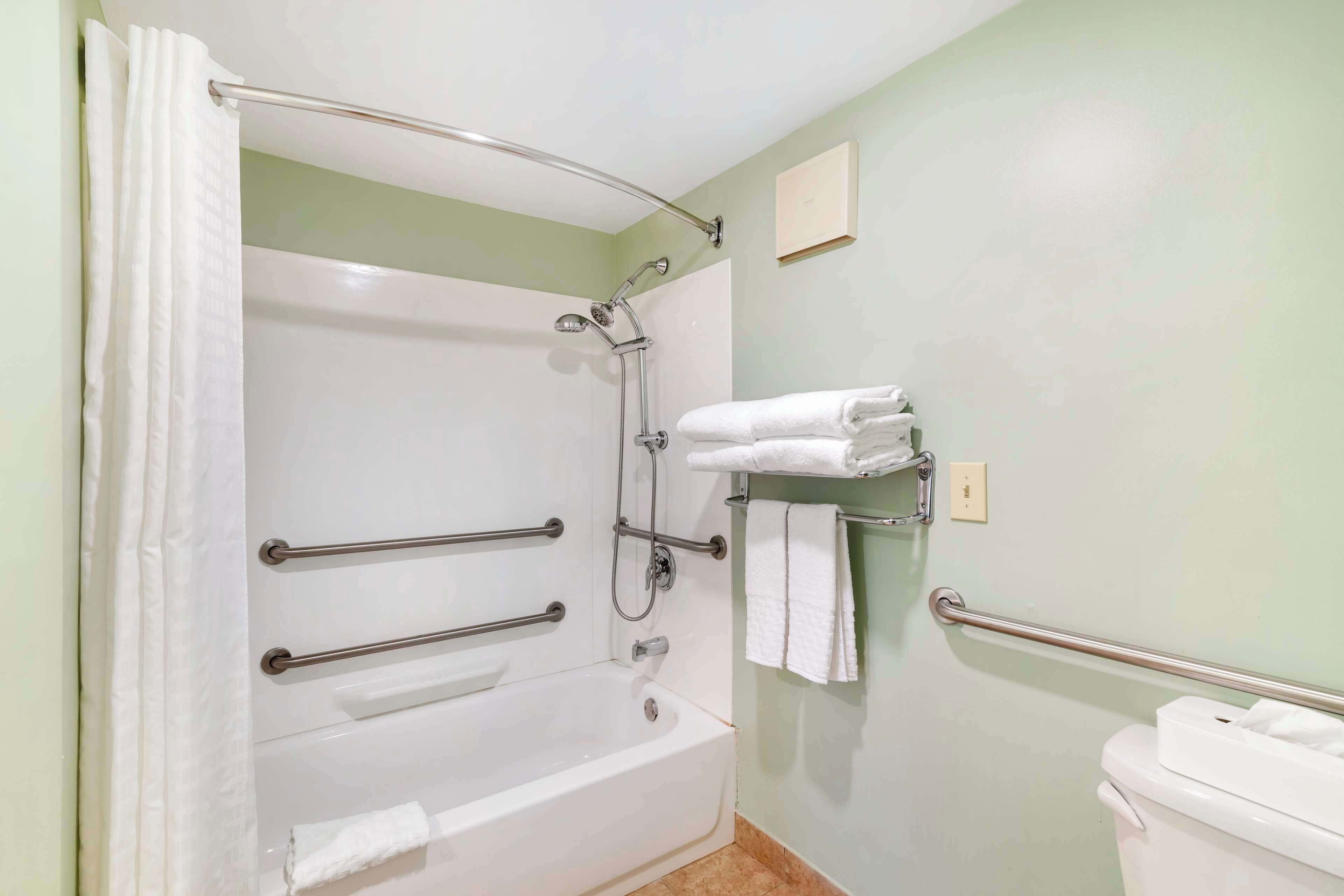 Un baño con bañera, ducha e inodoro. Cuenta con barras de apoyo para accesibilidad, un perchero y un interruptor de luz en la pared.