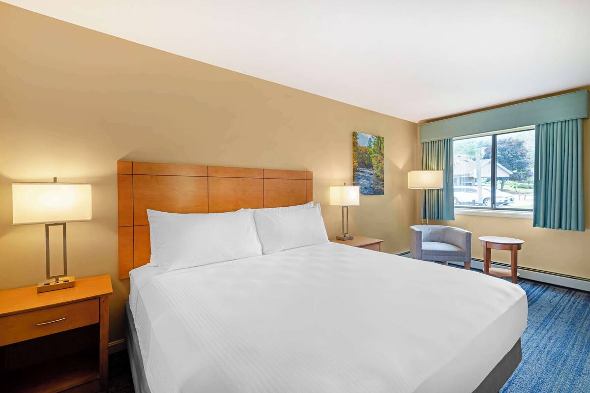 A hotel room with a large bed, two bedside lamps, a small table, a chair, and a window with curtains.