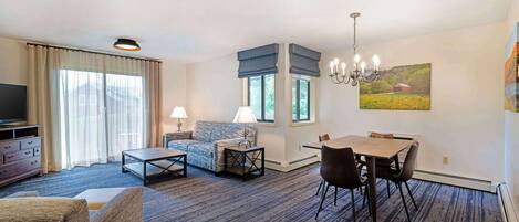 A hotel room with a sofa, a dining table with chairs, a TV, and a painting on the wall.