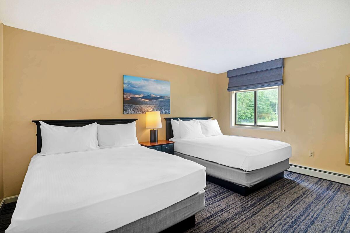 A hotel room with two beds, a nightstand with a lamp, a window with a view of greenery, and a painting on the wall.
