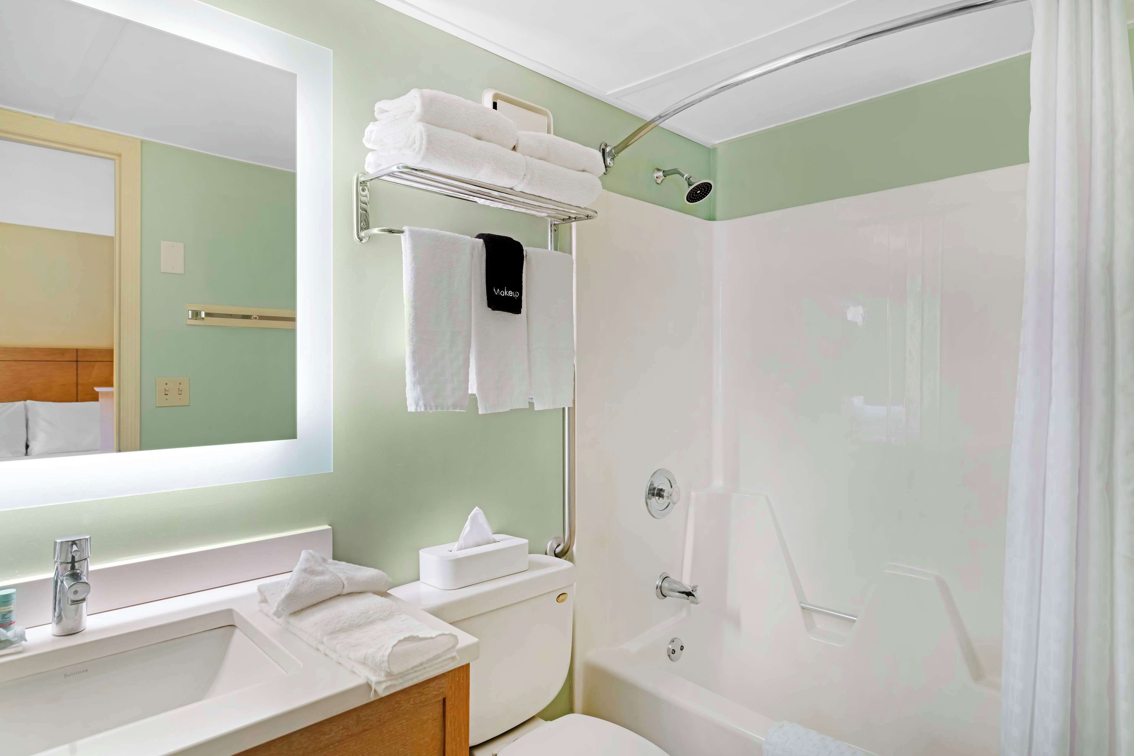 A bathroom with a sink, toilet, and shower. There are towels on the wall and a shelf with more towels.