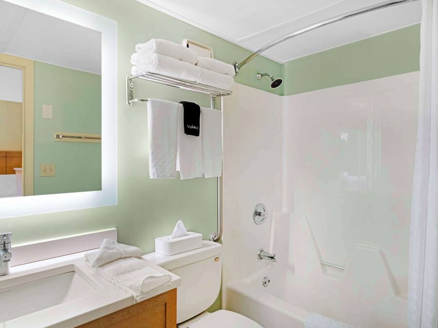 A bathroom with a sink, toilet, and shower. There are towels on the wall and a shelf with more towels.