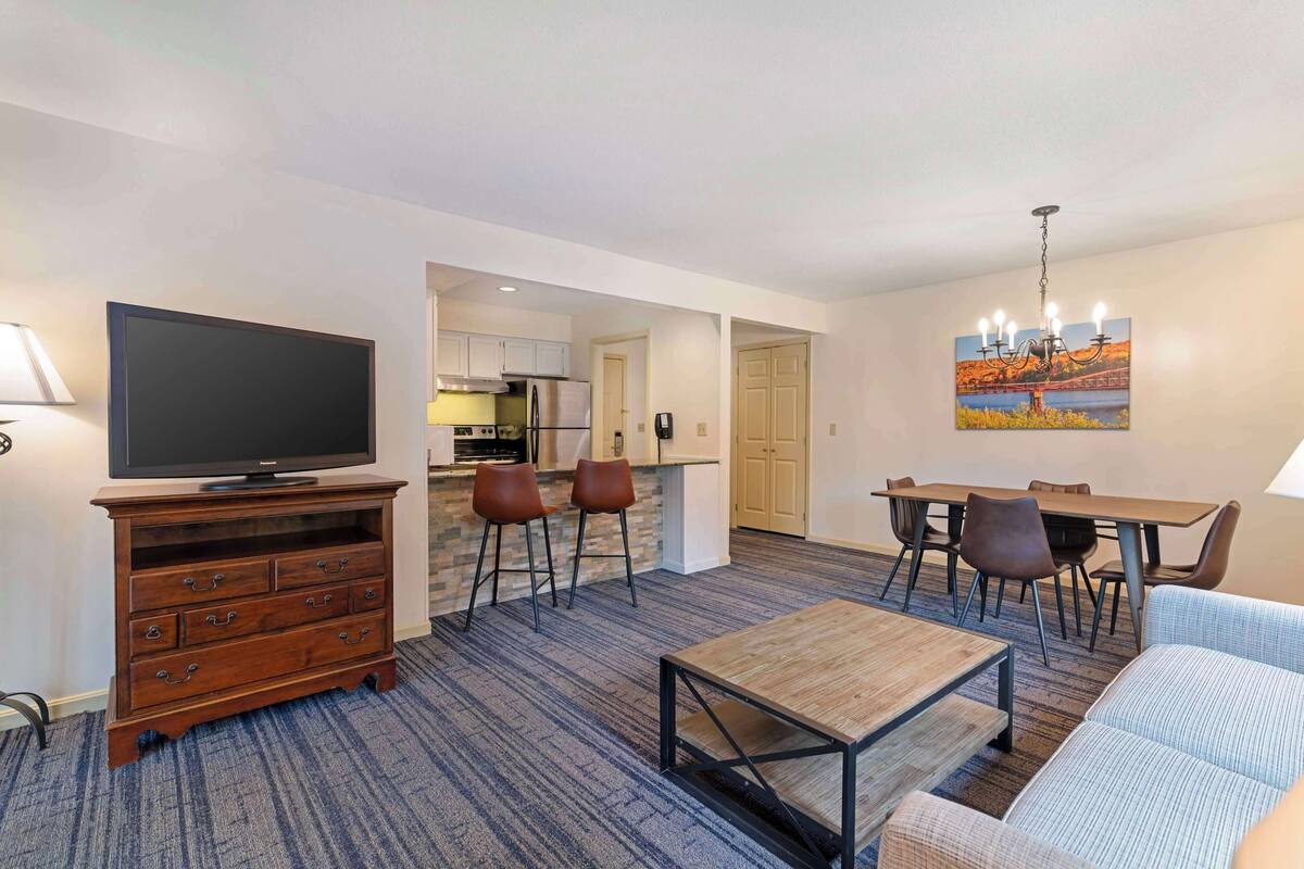 A hotel room with a TV, wooden dresser, sofa, dining table, chairs, and a kitchen area in the background.