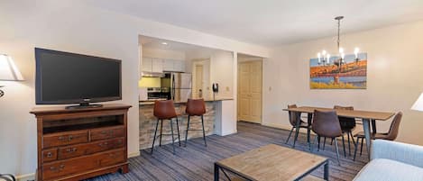 Quarto de hotel com televisão, cómoda de madeira, sofá, mesa de jantar, cadeiras e área de cozinha ao fundo.