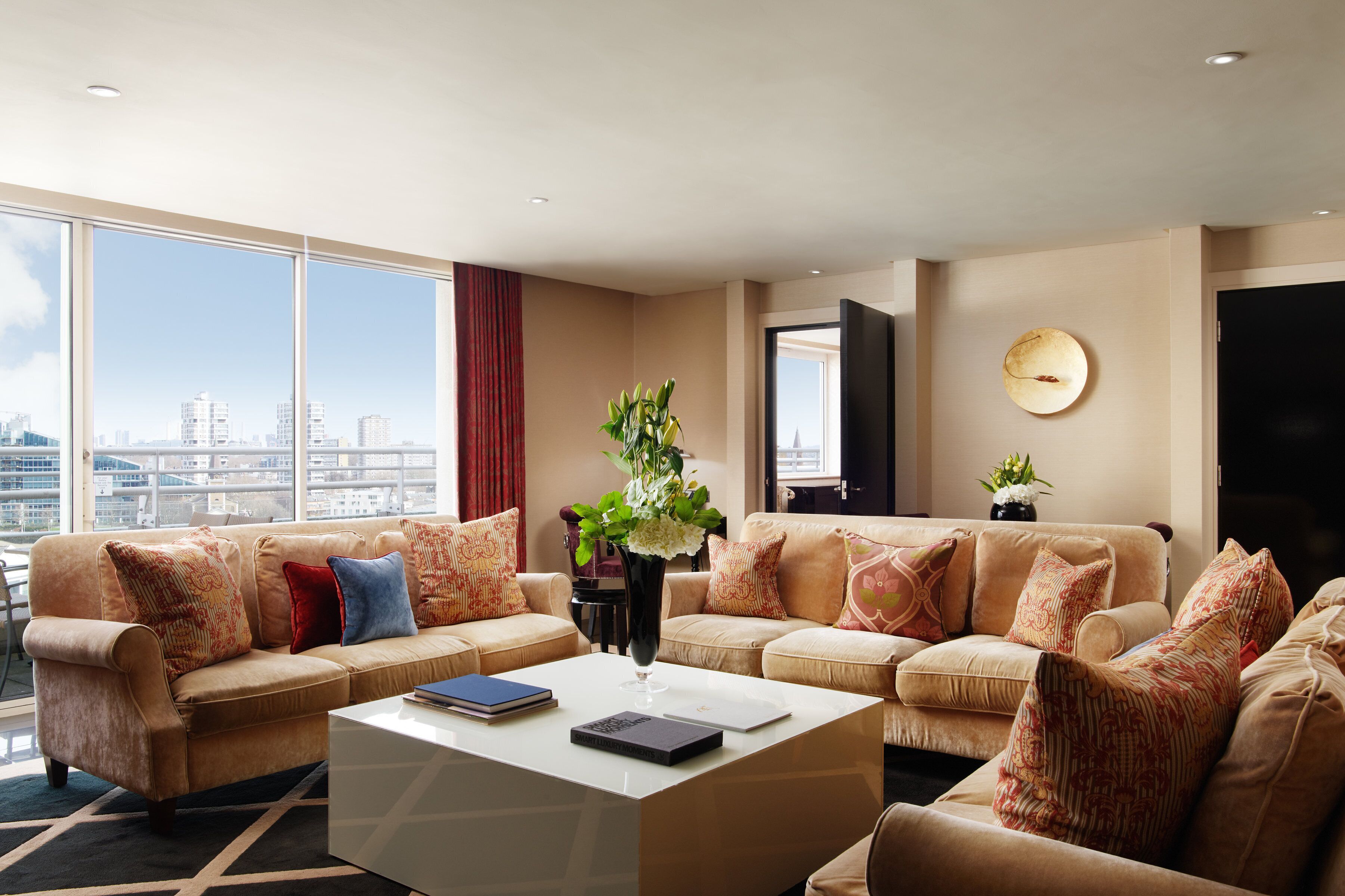 A spacious living room with a large window offering a city view, a white coffee table, and two beige sofas with patterned cushions.