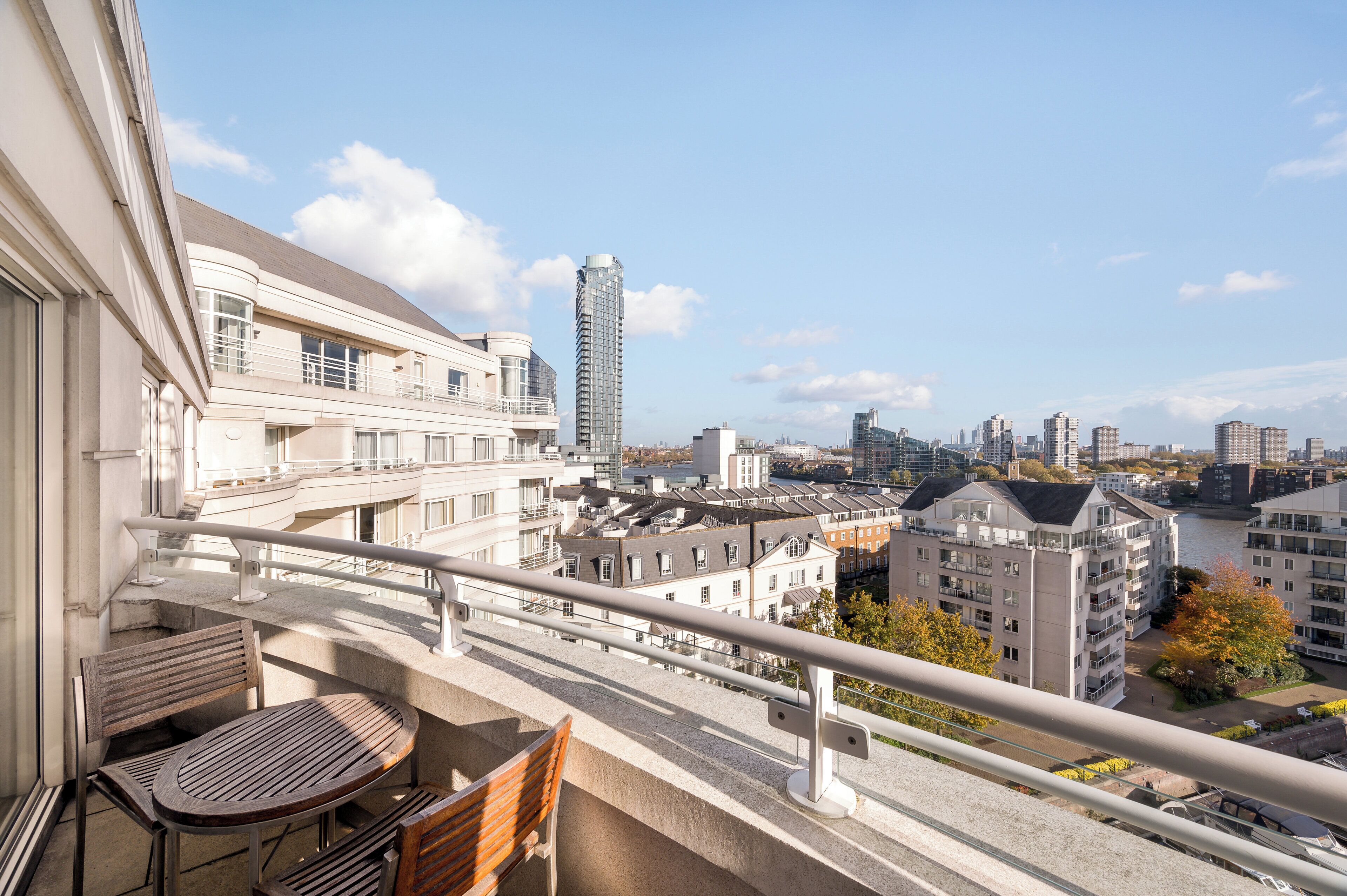 A balcony with a table and chairs, overlooking a cityscape with buildings and a river.
