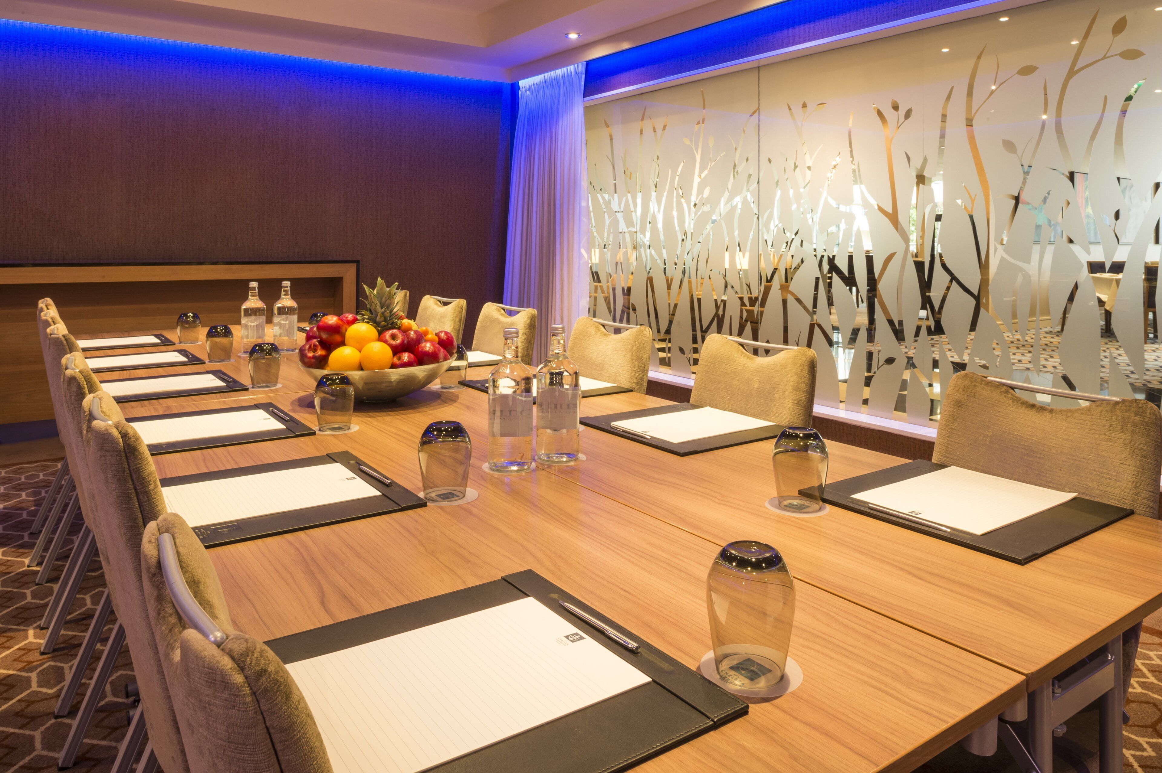 A conference room with a long table, chairs, a fruit bowl, water bottles, and a decorative wall.