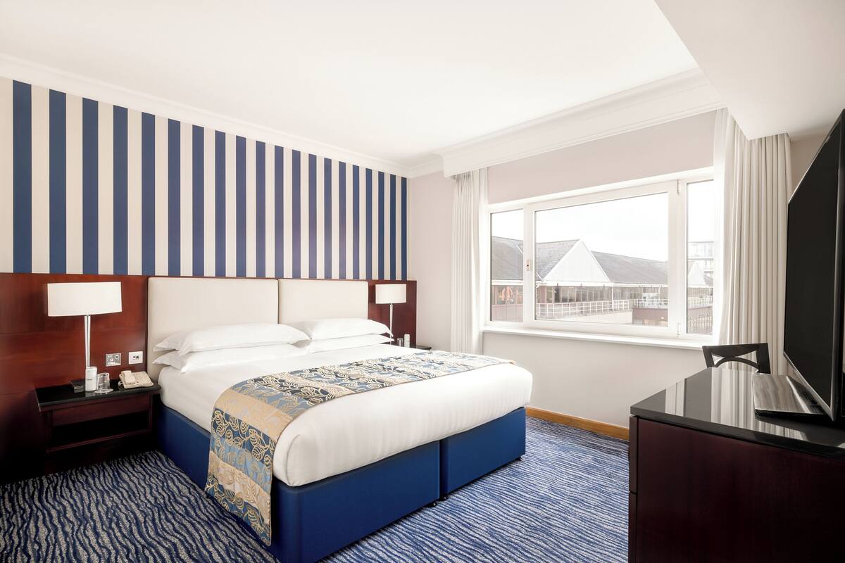 A hotel room with a large bed, a TV, a desk with a lamp, and a window with a view of buildings.