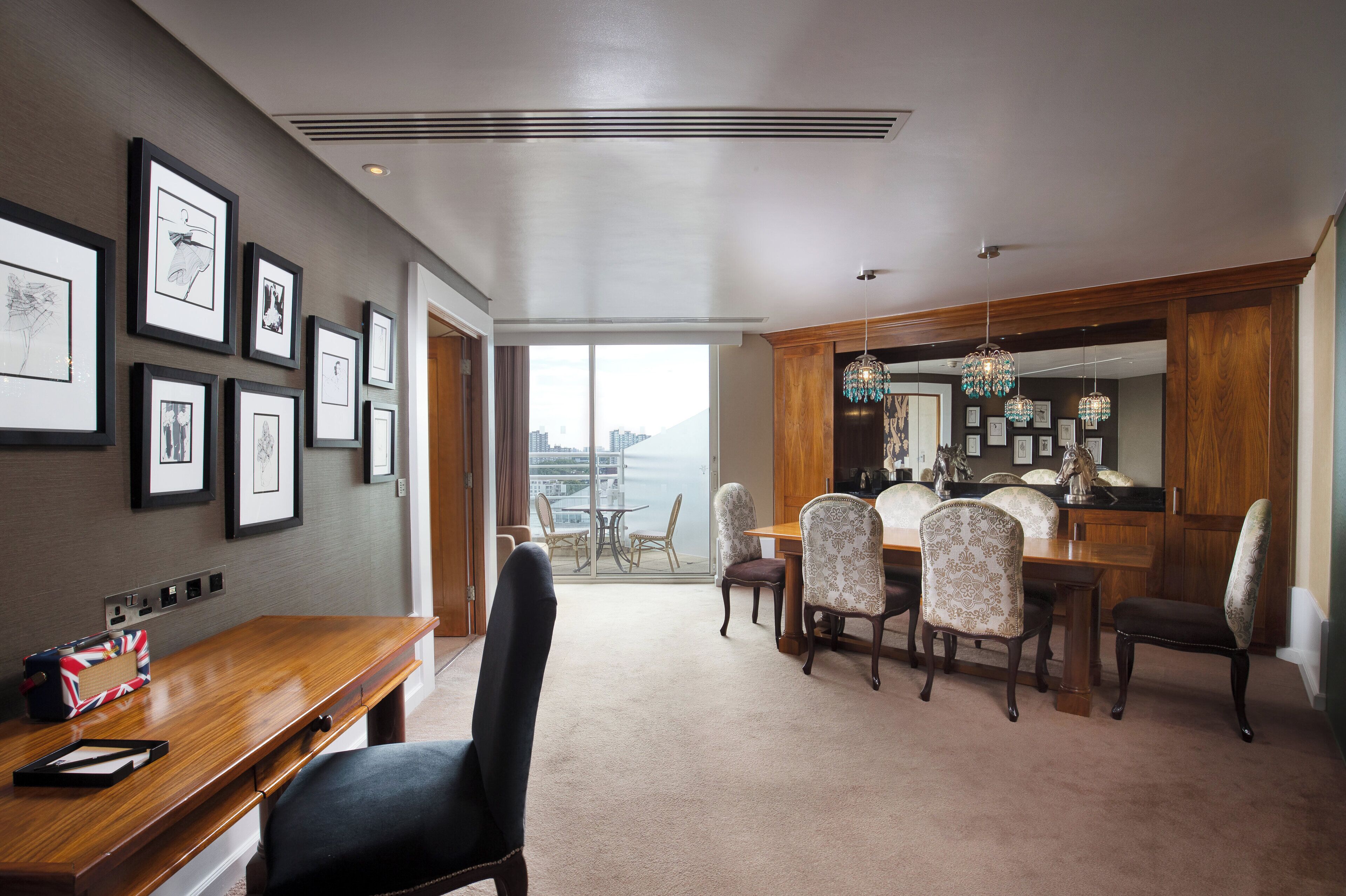 A hotel dining area with a wooden table and chairs, framed artwork on the walls, and a view through the glass door.
