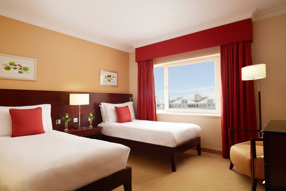 A hotel room with two beds, a desk, a chair, a window with red curtains, and artwork on the walls.