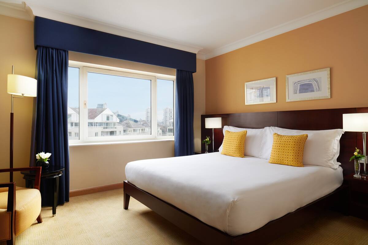 A hotel room with a large bed, two bedside lamps, a nightstand, a chair, a window with curtains, and framed pictures on the wall.