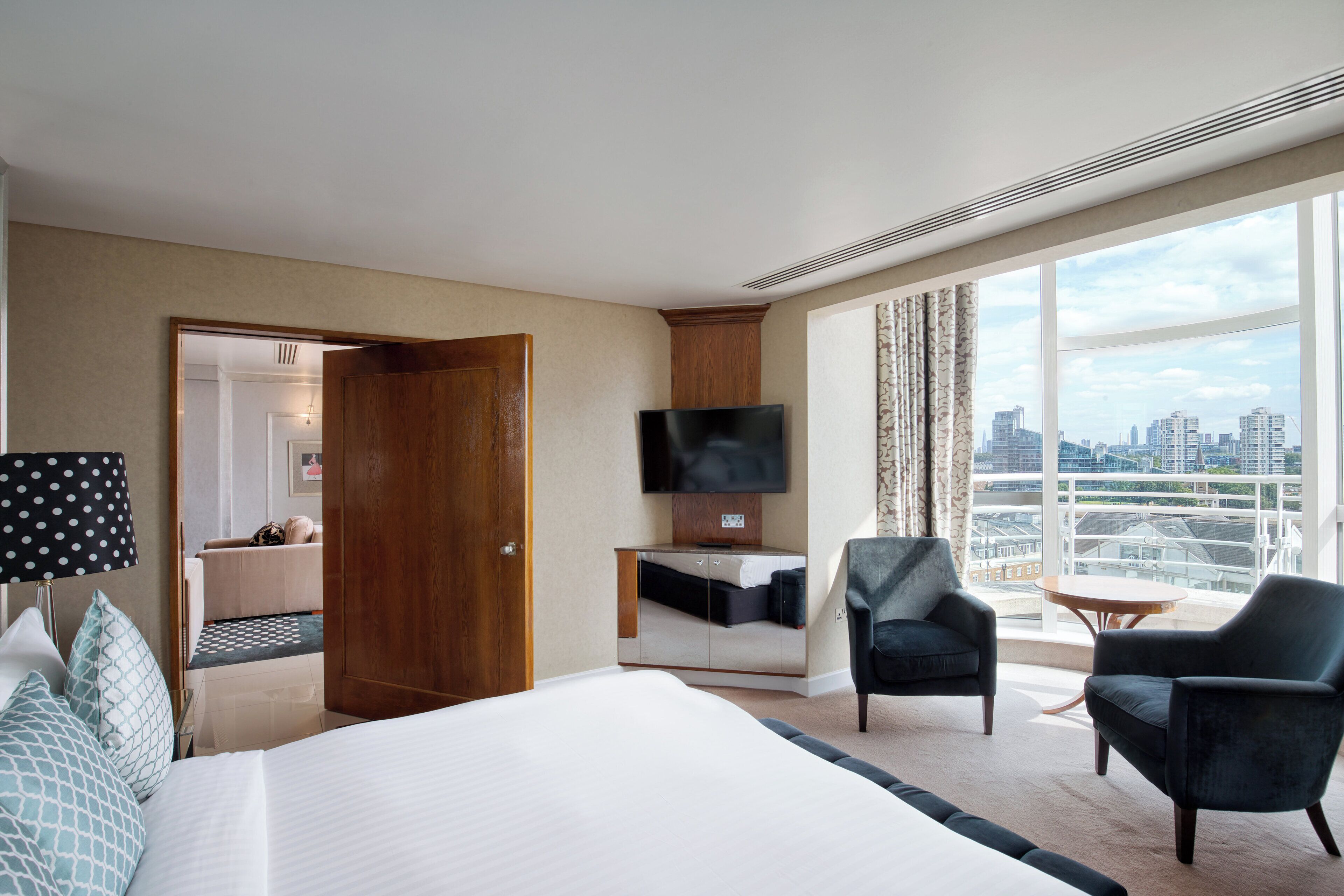 A hotel room with a large bed, a desk with a flat-screen TV, two armchairs, a small table, and a city view.