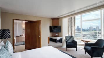 A hotel room with a large bed, a desk with a flat-screen TV, two armchairs, a small table, and a city view.