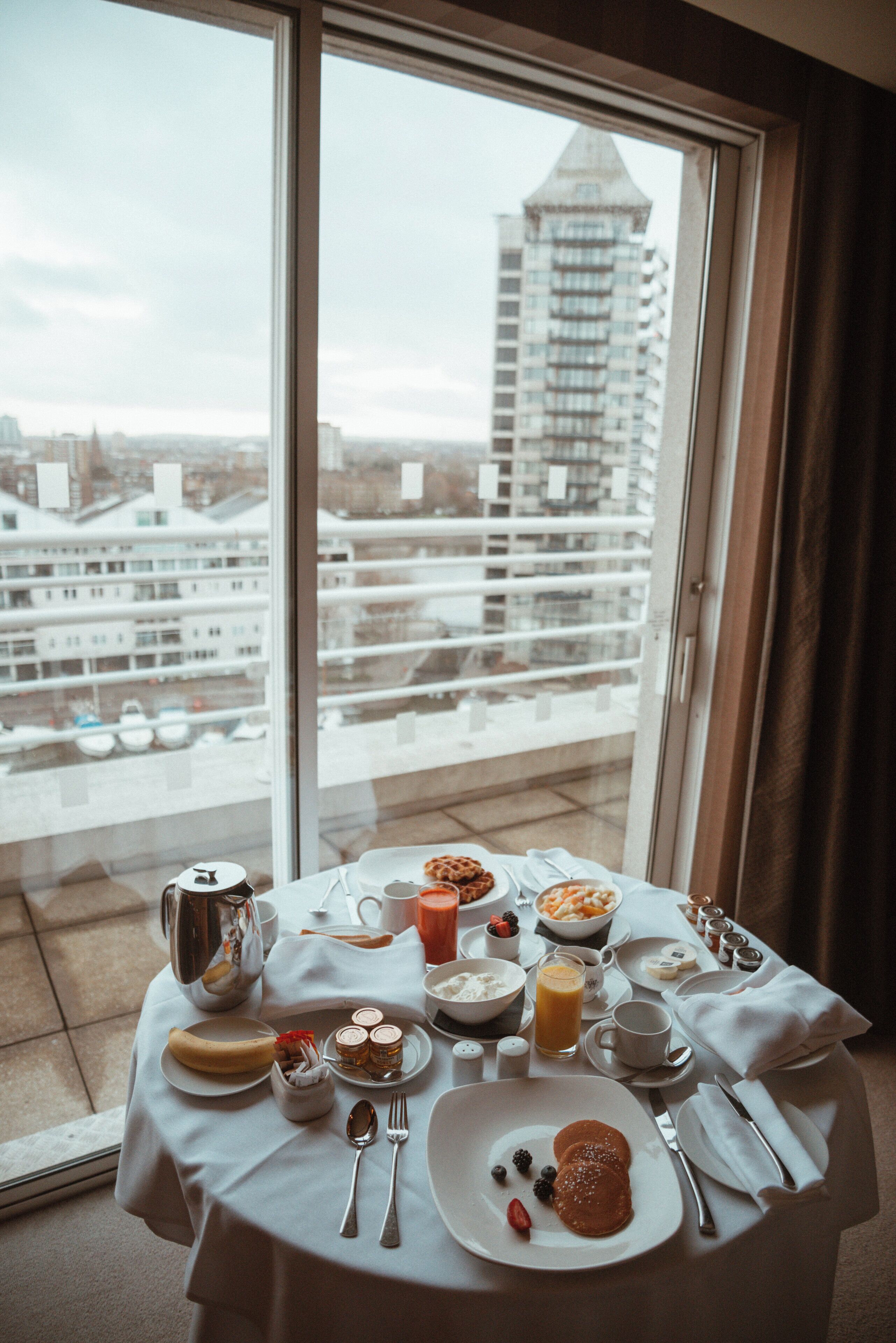 A breakfast table with various food items and utensils, set on a balcony with a city view.