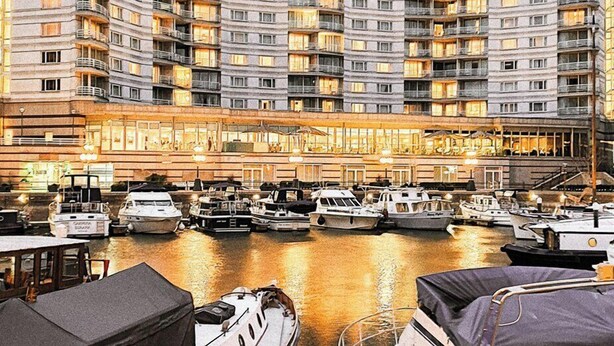 Un port de plaisance où sont amarrés plusieurs bateaux, un grand bâtiment d’hôtel doté de nombreux balcons et de fenêtres illuminées, et une zone de quai en bois.