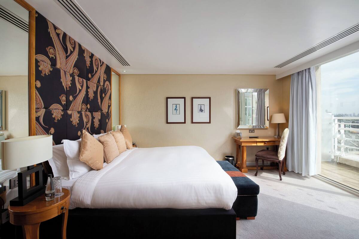 A hotel room with a large bed, a desk, and a chair. There are two framed pictures on the wall, a window with curtains, and a view of the city.