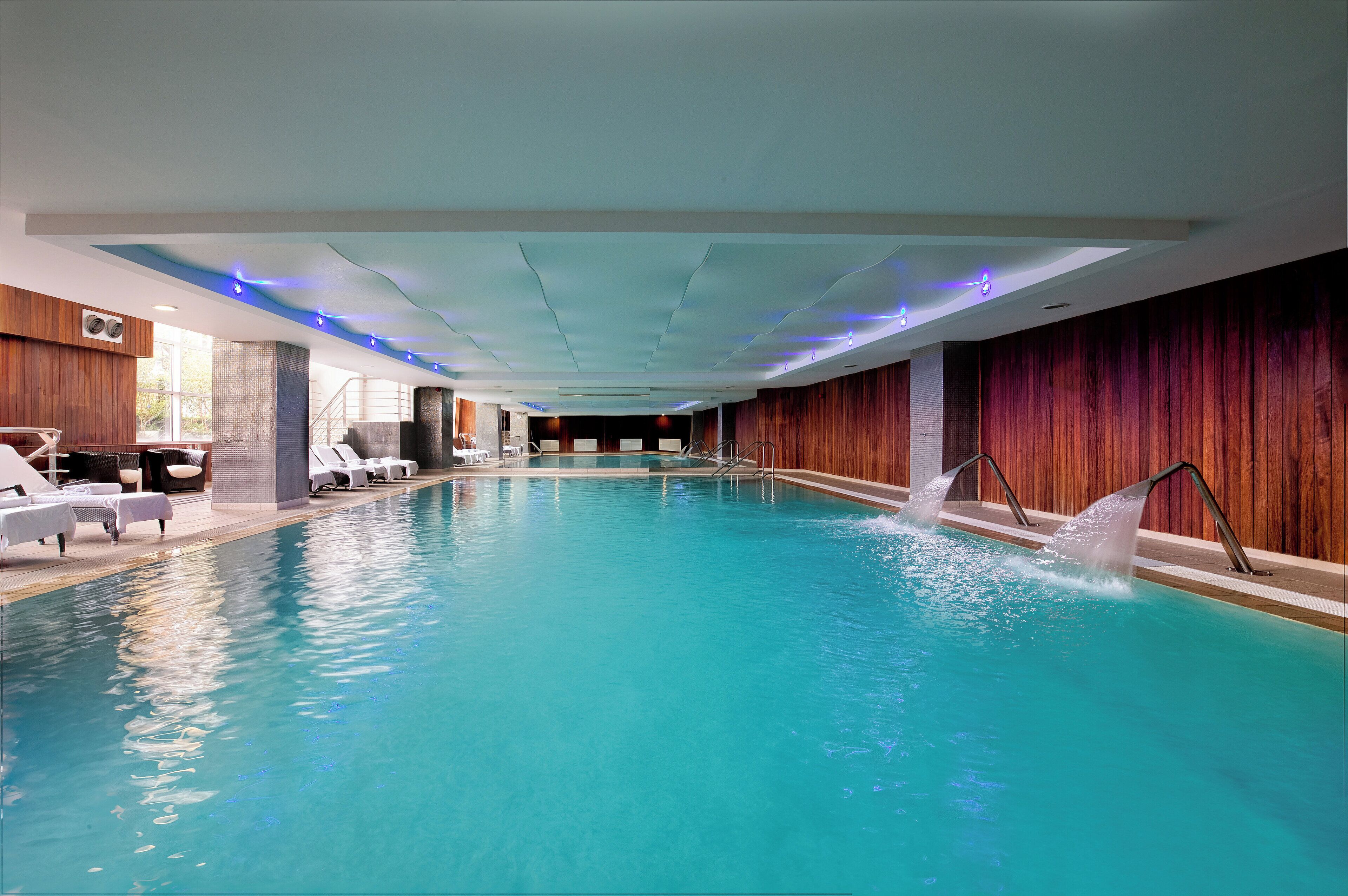 An indoor swimming pool with clear blue water, a wooden wall on one side, and a seating area with white lounge chairs and a table.