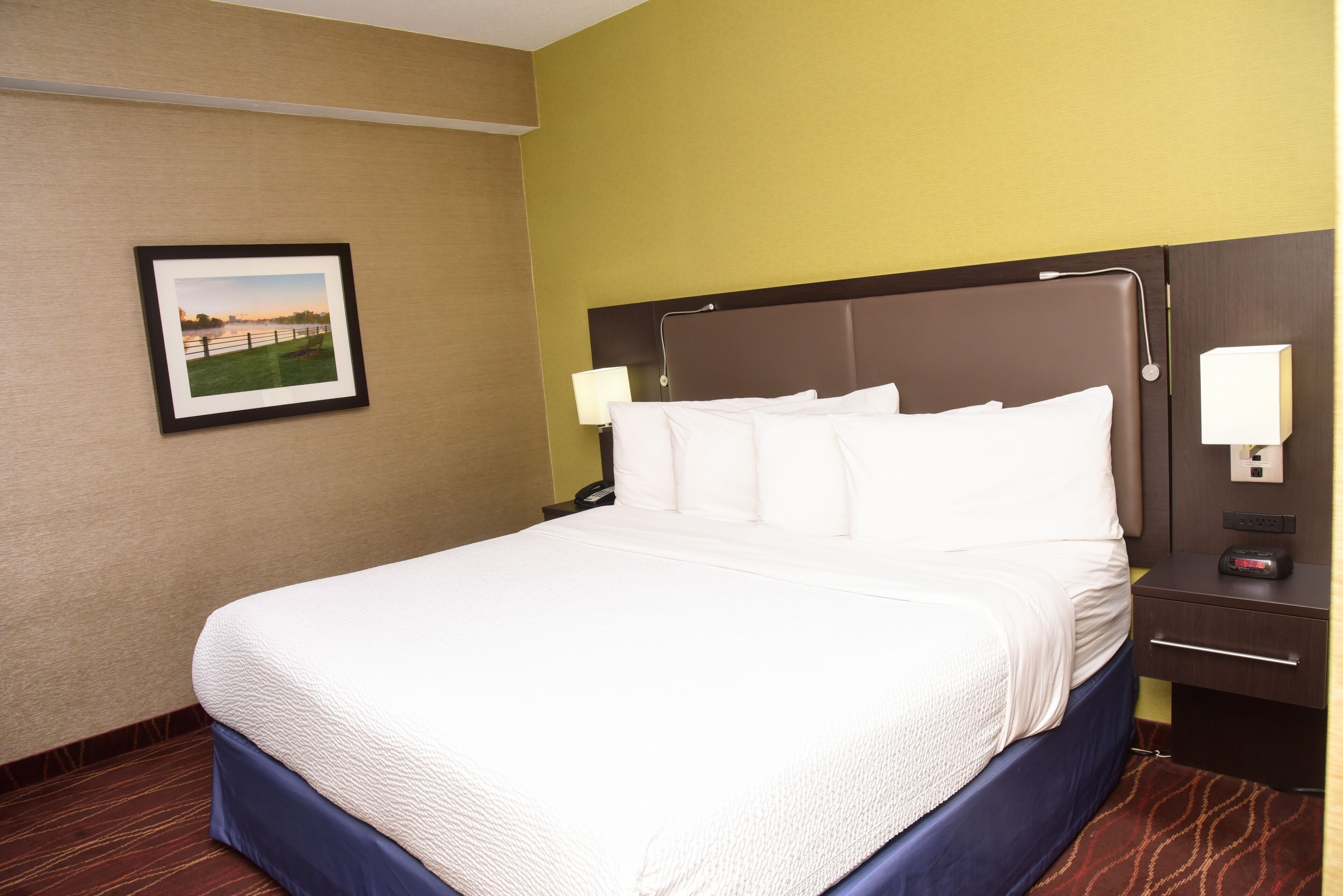Een hotelkamer met een groot bed, een nachtkastje met een lamp, een lijst aan de muur en een bruin hoofdeinde.