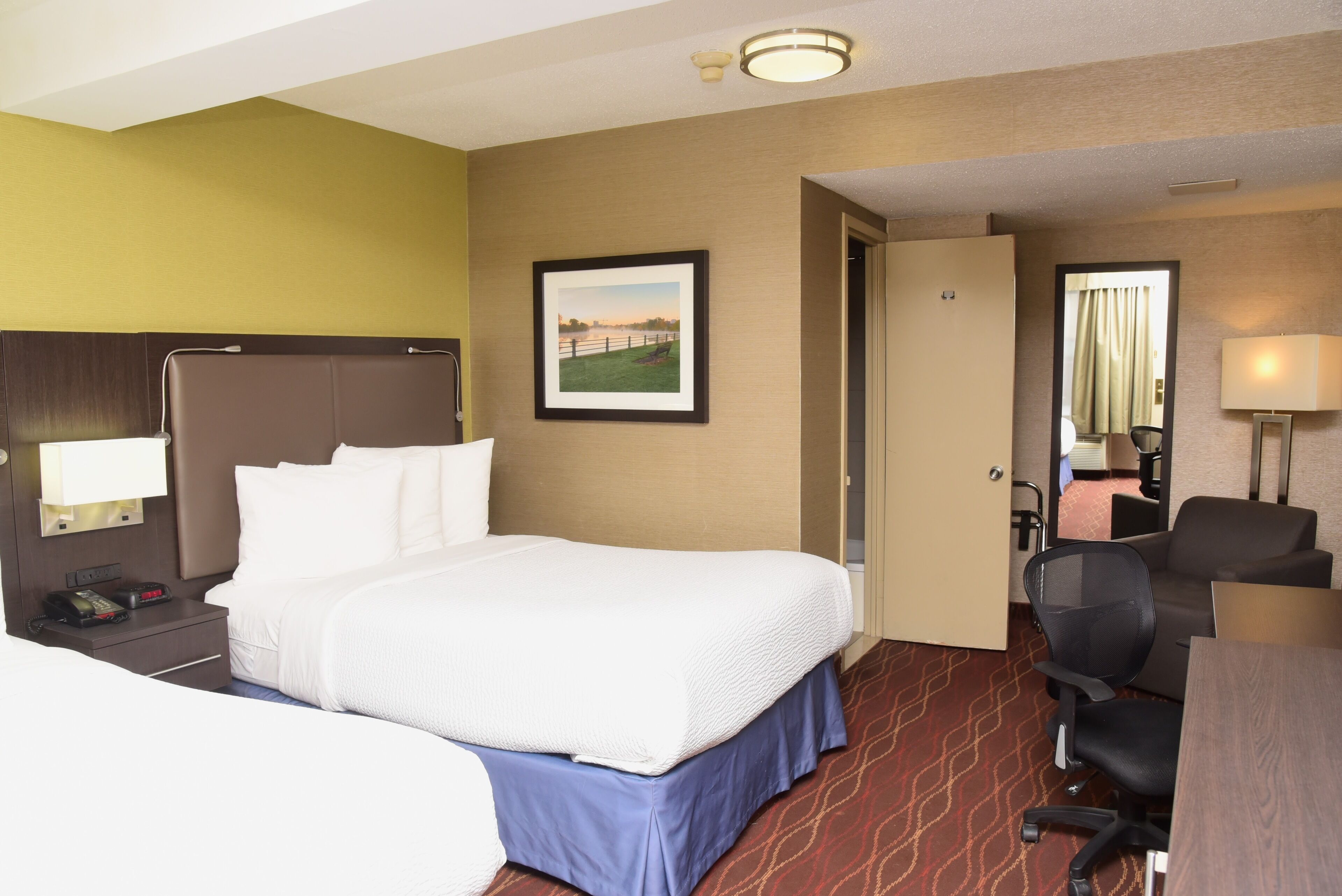 A hotel room with a bed, a desk, a chair, a nightstand with a phone, and a picture on the wall.