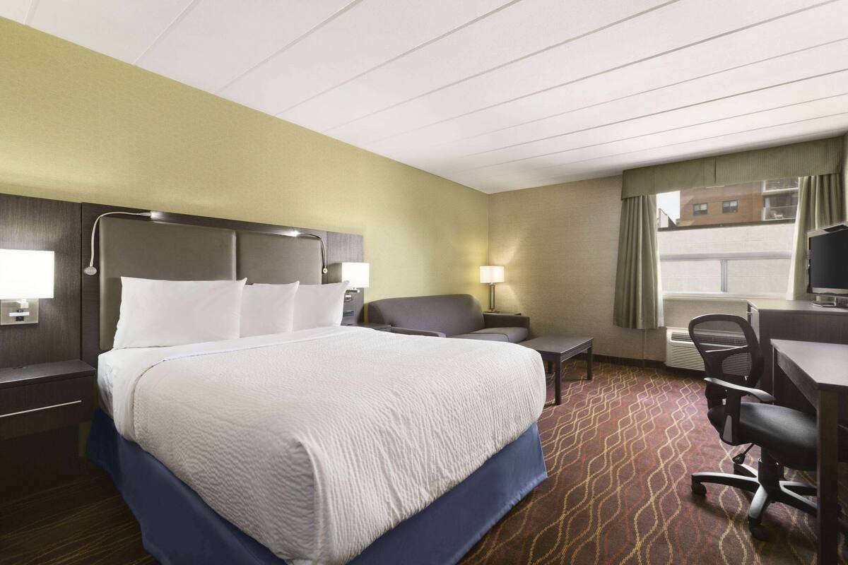 A hotel room with a large bed, a desk, a chair, a lamp, and a view of buildings through the window.