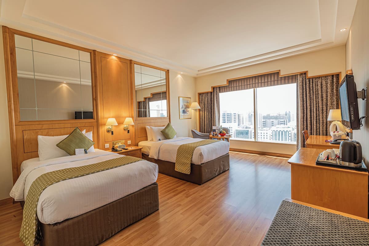 Een hotelkamer met twee bedden, een bureau, een stoel, een groot raam met uitzicht op de stad en een spiegel.